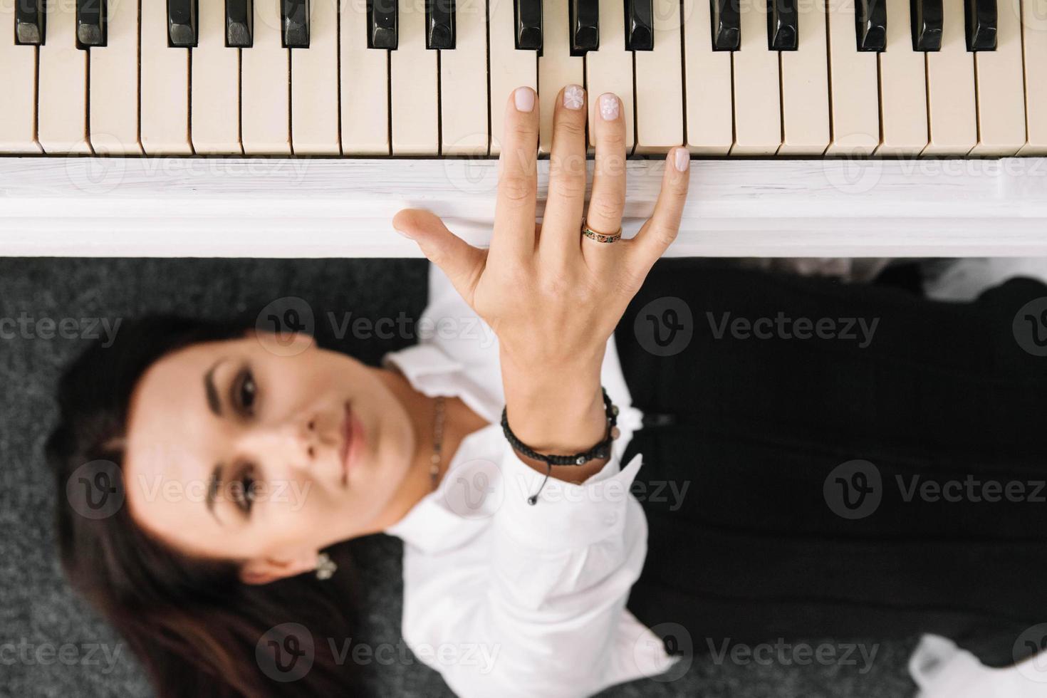 linda mulher vestida com um vestido branco com um espartilho preto encontra-se no chão perto de um piano branco tocando as teclas. lugar para texto ou publicidade. vista de cima foto