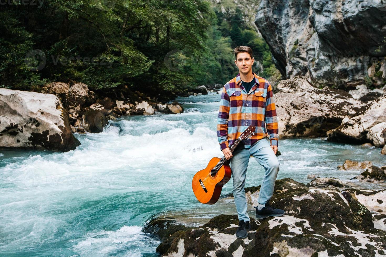 jovem segurando uma guitarra em pé na margem de um rio de montanha em um fundo de rochas e floresta foto