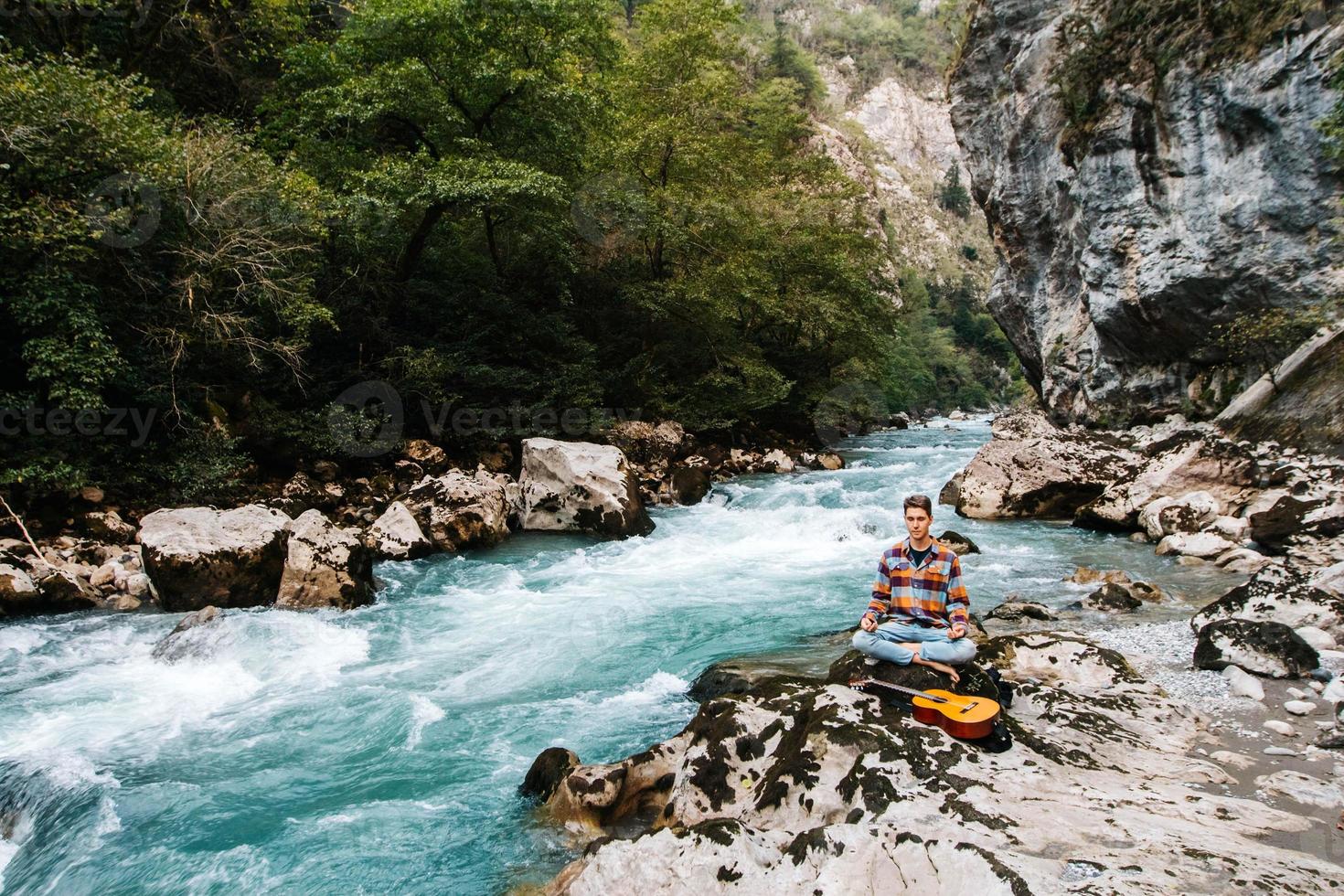 homem em posição meditativa com guitarra sentado na margem de um rio de montanha em um fundo de rochas e floresta foto