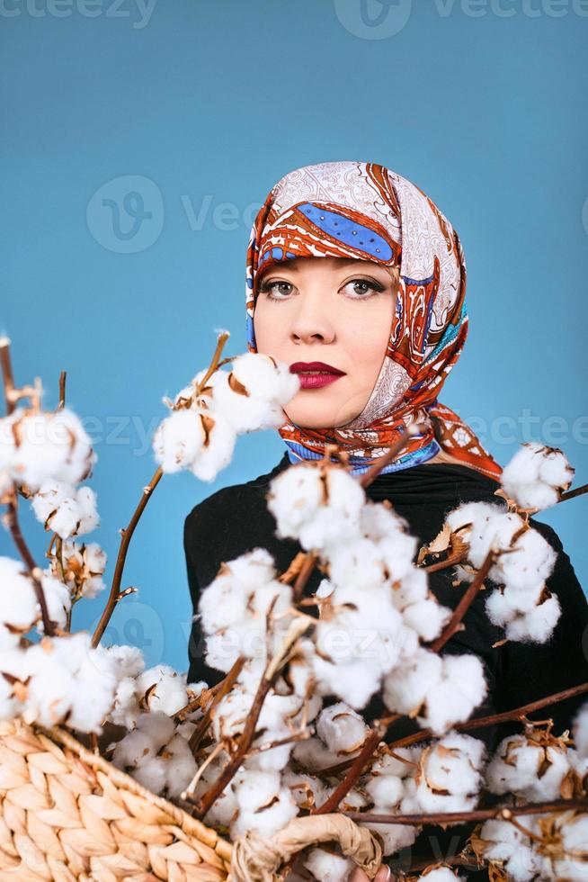 mulher oriental em lenço colorido, segurando a cesta com galhos de algodão sobre fundo azul. coletor de algodão foto