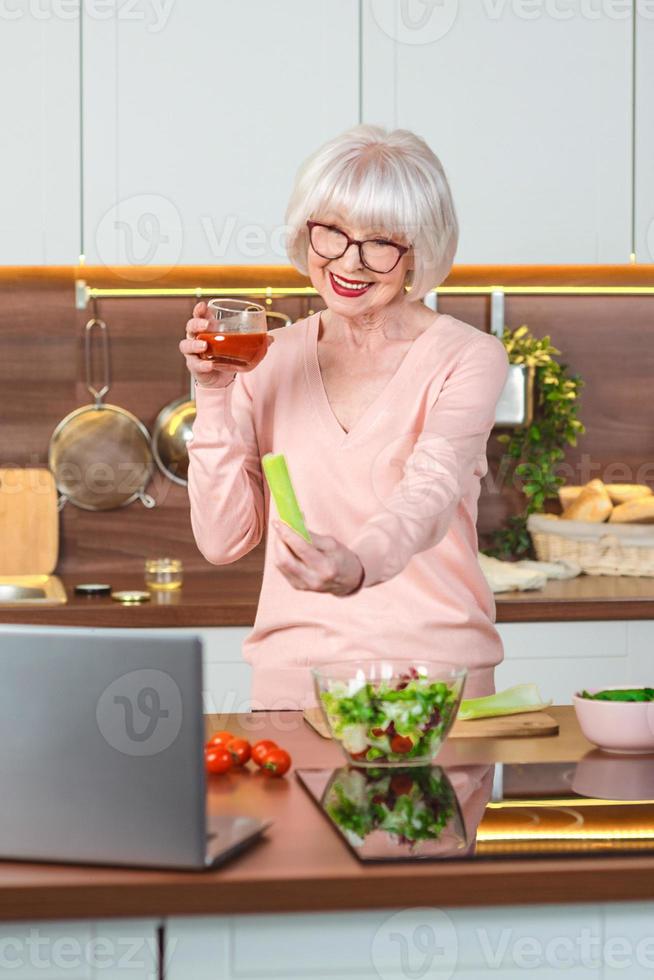 blogueira de vídeo sênior de mulher alegre, mostrando suco de aipo e tomate em sua cozinha. cru, vegetariano, dieta, blogueiro, conceito de cozinha nutricionista foto