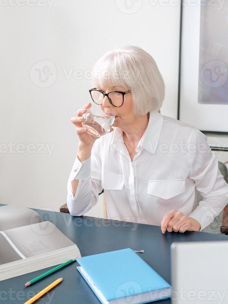 sênior linda mulher de cabelos grisalhos na blusa branca, bebendo água durante o trabalho no escritório. trabalho, idosos, equilíbrio hídrico, encontrar uma solução, conceito de experiência foto