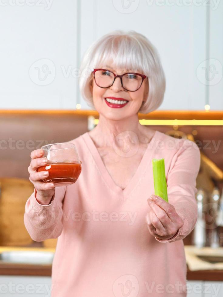 mulher alegre sênior com suco de aipo e tomate na dieta em sua cozinha. cru, vegetariano, dieta, conceito de cozinha foto