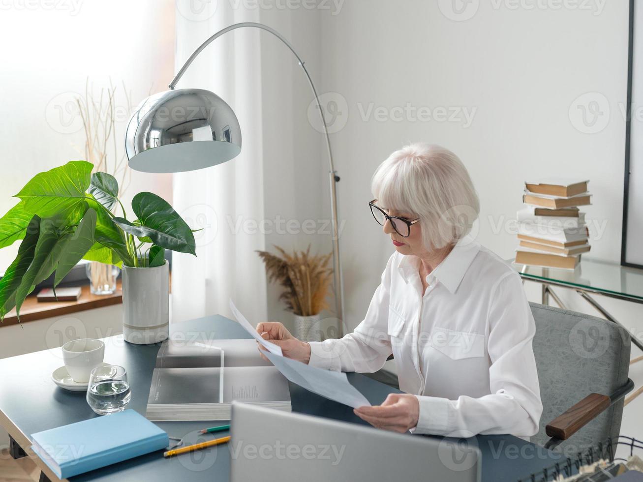 cansado sênior linda mulher de cabelo grisalho na blusa branca, lendo documentos no escritório. trabalho, pessoas seniores, problemas, encontrar uma solução, conceito de experiência foto