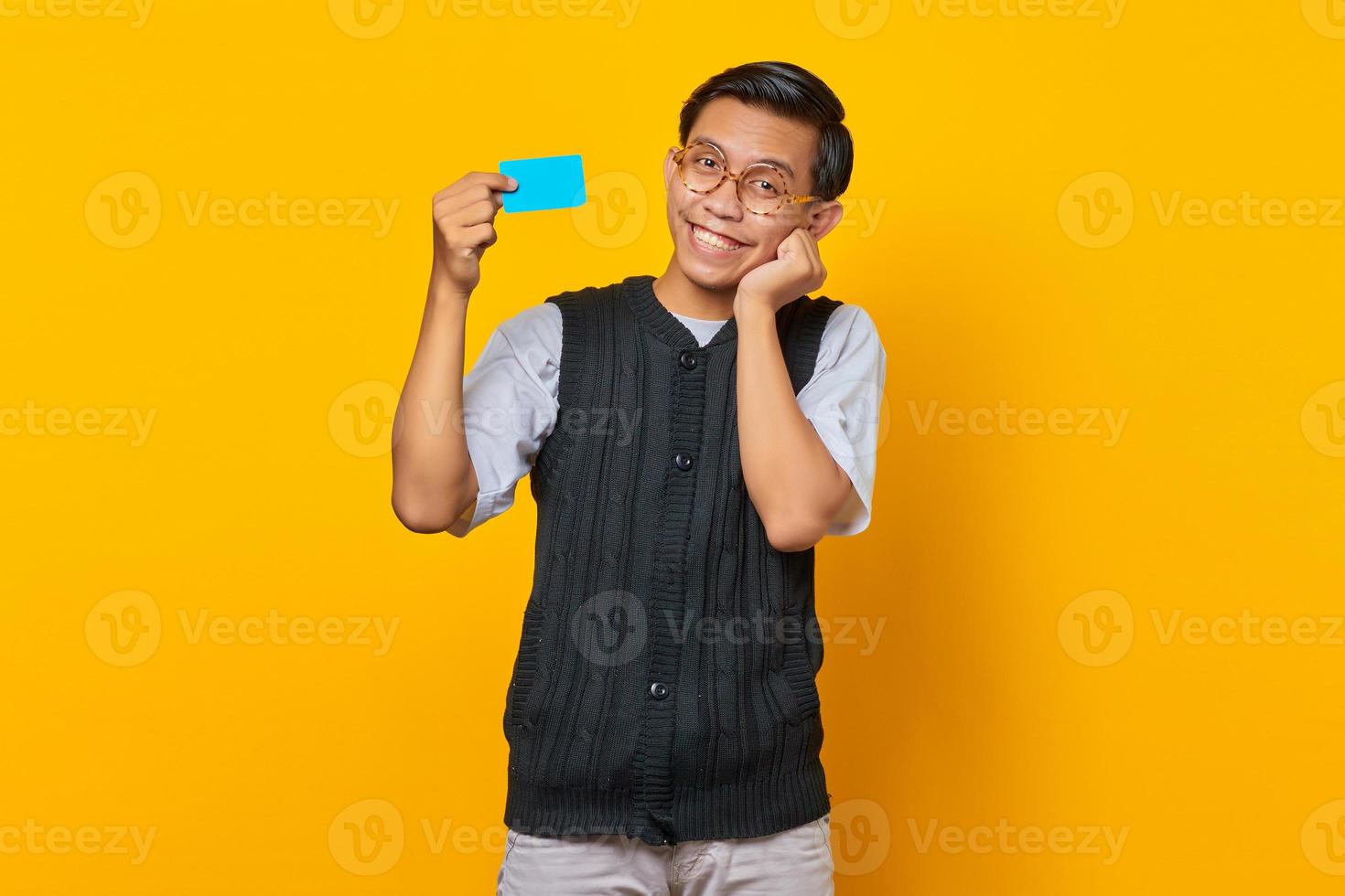 homem asiático sorridente segurando um cartão de crédito com a boca aberta e as mãos nas bochechas em fundo amarelo foto