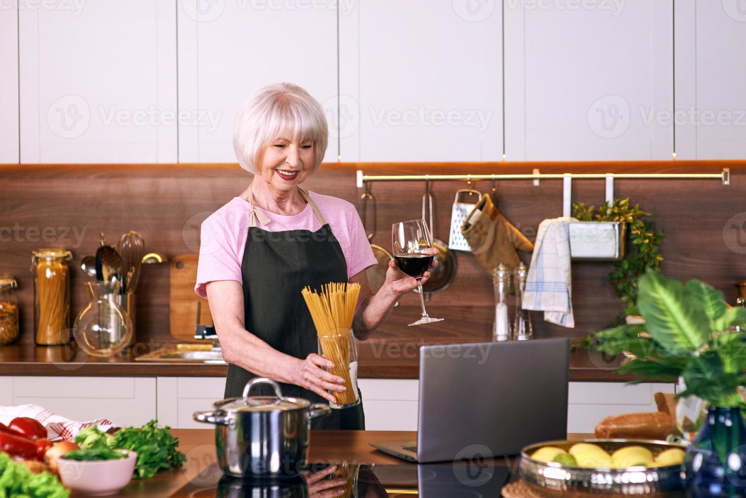mulher alegre sênior está cozinhando na cozinha moderna, por laptop. comida, educação, conceito de estilo de vida foto