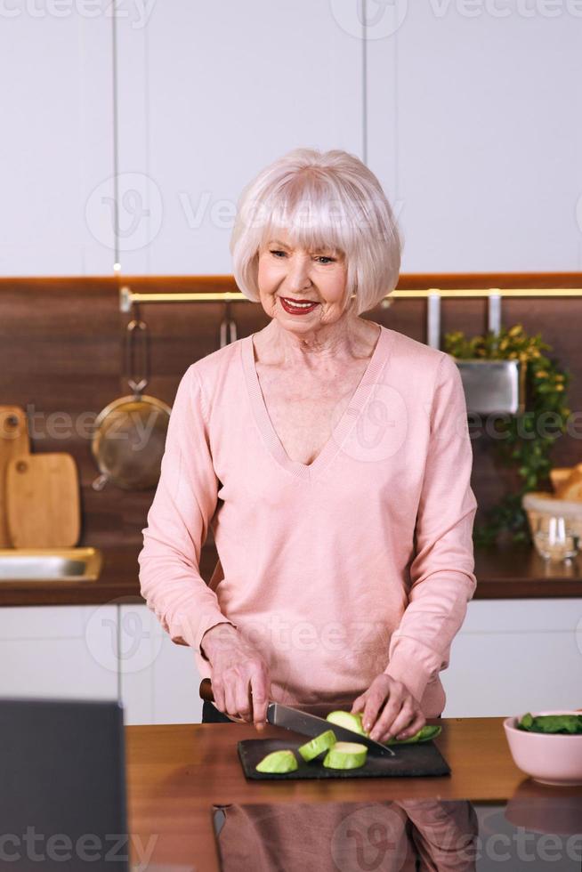 mulher alegre sênior está cozinhando na cozinha moderna, por laptop. comida, educação, conceito de estilo de vida foto
