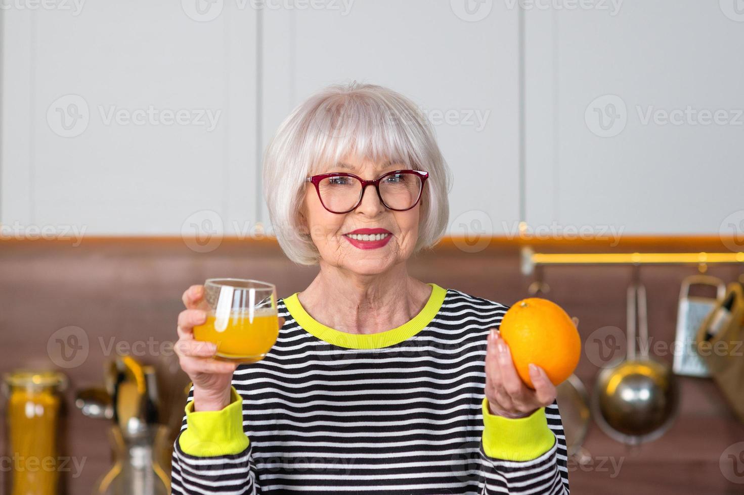 alegre bonita sênior mulher sorridente de suéter listrado, bebendo suco de laranja em pé na cozinha. saudável e suculento estilo de vida, casa, conceito de pessoas sênior. foto