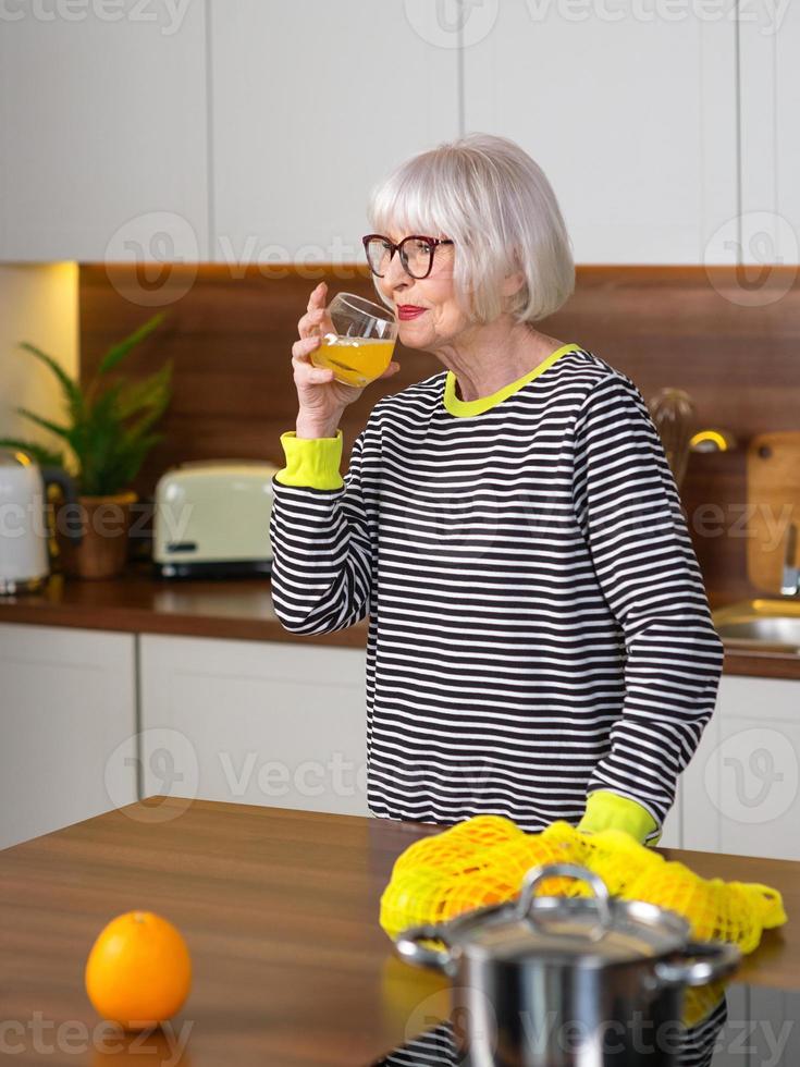 alegre bonita sênior mulher sorridente de suéter listrado, bebendo suco de laranja em pé na cozinha. saudável e suculento estilo de vida, casa, conceito de pessoas sênior. foto