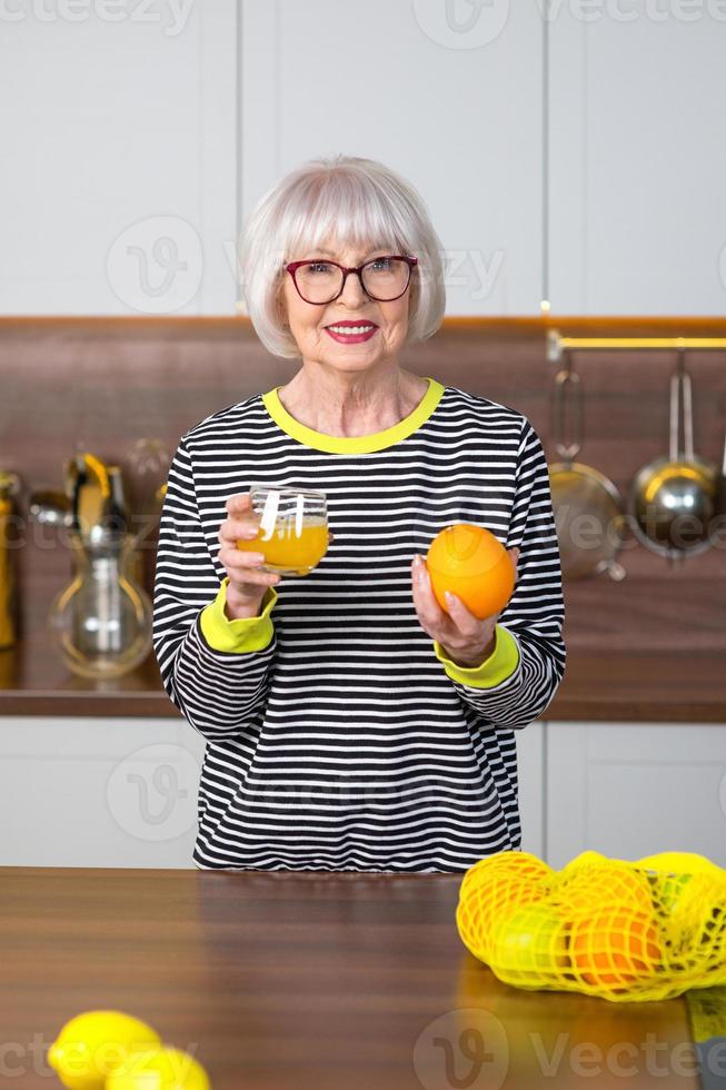 alegre bonita sênior mulher sorridente de suéter listrado, bebendo suco de laranja em pé na cozinha. saudável e suculento estilo de vida, casa, conceito de pessoas sênior. foto