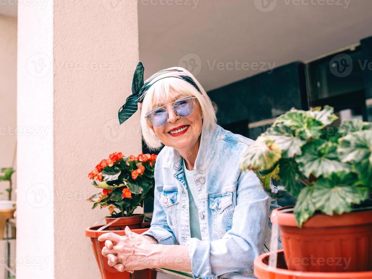 mulher elegante sênior com cabelos grisalhos e óculos azuis e jeans em pé na varanda ao ar livre. foto