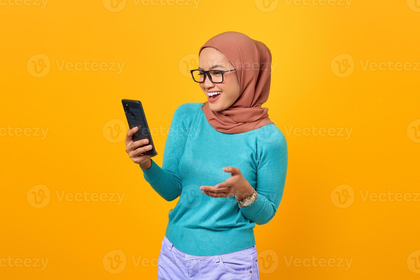 Mulher asiática jovem animada usando smartphone e comemorando o sucesso isolado em fundo amarelo foto