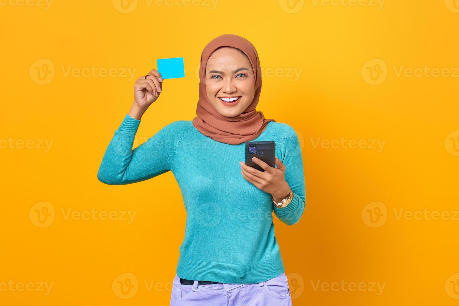 jovem mulher asiática sorridente, mostrando o cartão de crédito e segurando um telefone celular no fundo amarelo foto