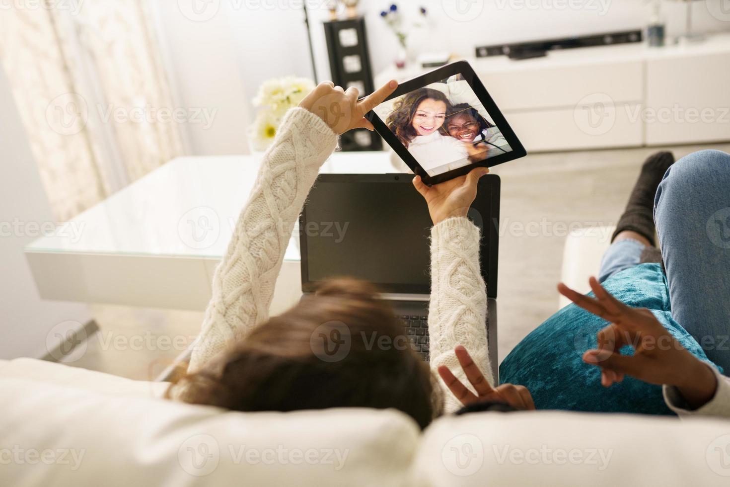 duas amigas fazendo uma selfie com um tablet digital, sentadas no sofá em casa. foto