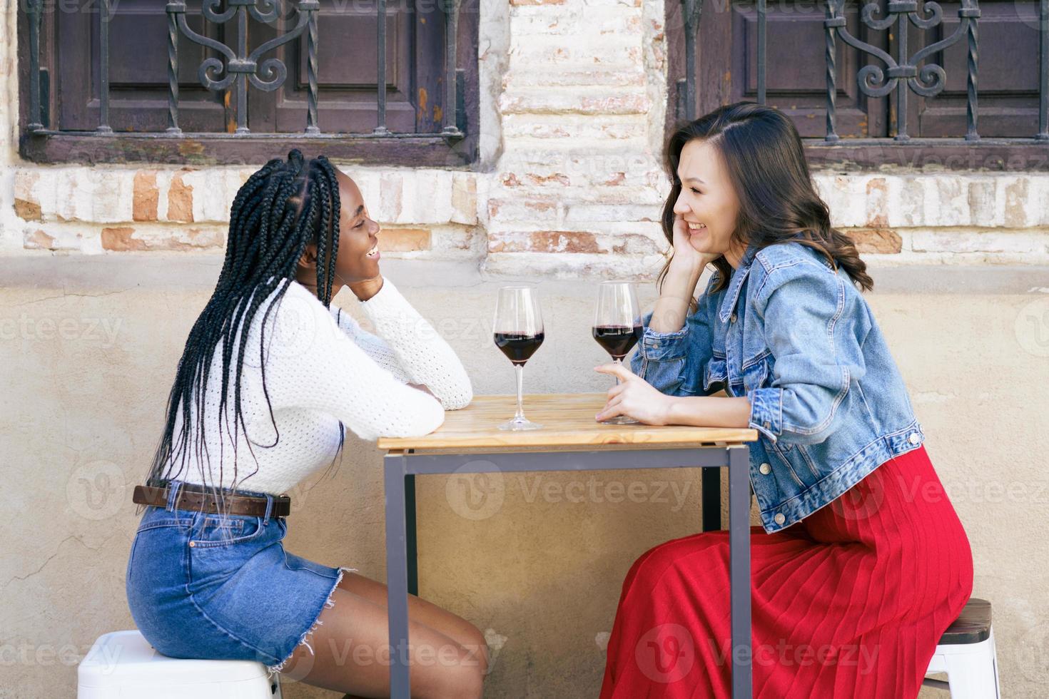 dois amigos multiétnicos conversando, sentados à mesa do lado de fora de um bar, enquanto bebem uma taça de vinho tinto. foto