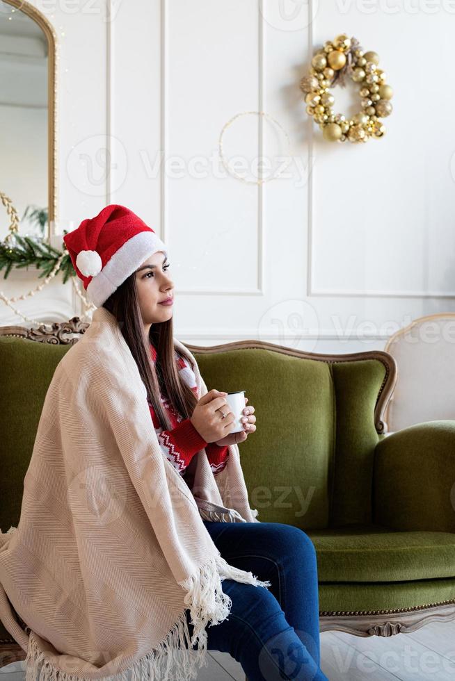 jovem sentada sozinha no sofá em uma sala de estar decorada para o natal tomando uma bebida quente foto