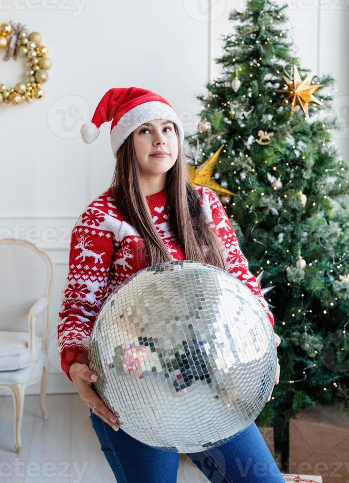 jovem com chapéu de Papai Noel segurando uma bola de espelhos em pé ao lado da árvore de Natal foto