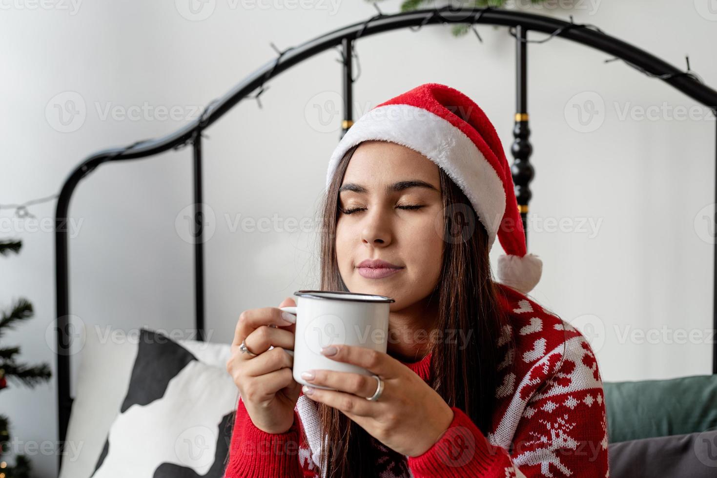 jovem de suéter vermelho e chapéu de Papai Noel sentada na cama comemorando o natal tomando uma bebida quente foto