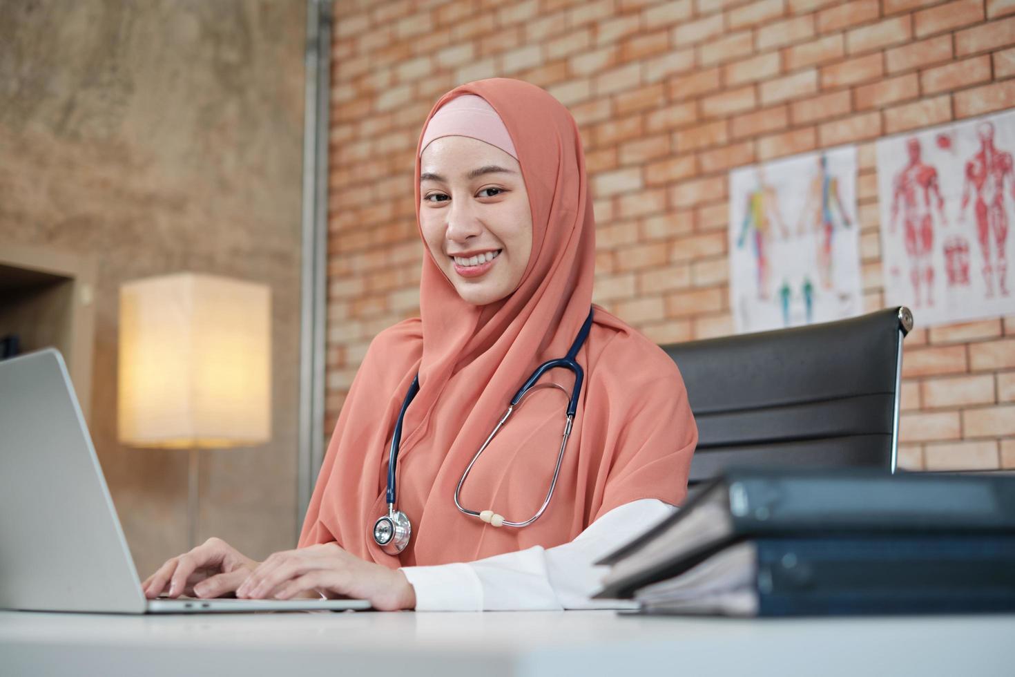 retrato de uma linda médica, linda muçulmana de uniforme com um estetoscópio, sorrindo e trabalhando com o laptop na clínica do hospital. uma pessoa com experiência em tratamento profissional. foto