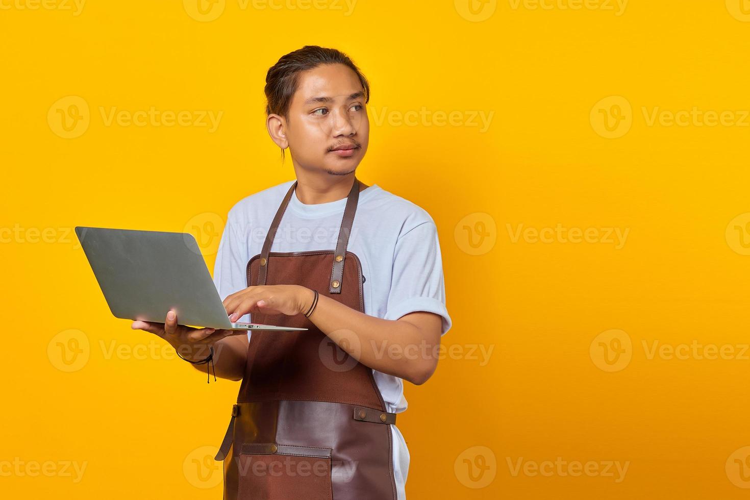 alegre e bonito jovem barista segurando um laptop e olhando de soslaio sobre fundo amarelo foto