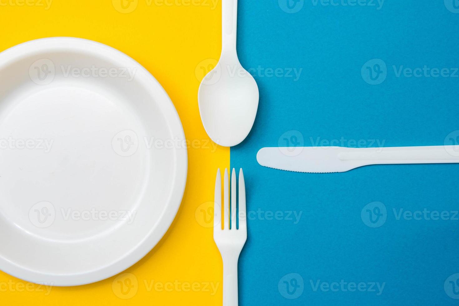 garfo, faca, colher e prato de plástico branco sobre fundo amarelo e azul foto