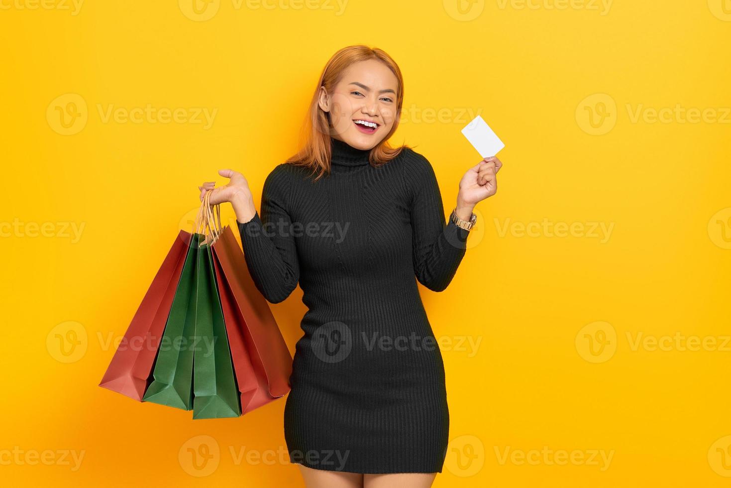 jovem mulher asiática sorridente segurando sacolas de compras e um cartão plástico isolado sobre fundo amarelo foto