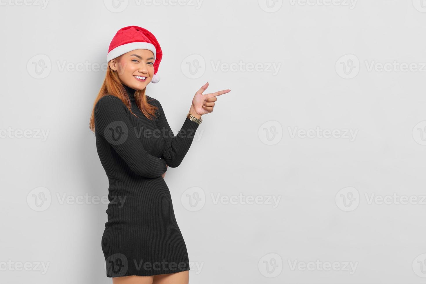 Mulher asiática jovem sorridente com chapéu de Papai Noel apontando o dedo para o espaço da cópia isolado sobre o fundo branco foto