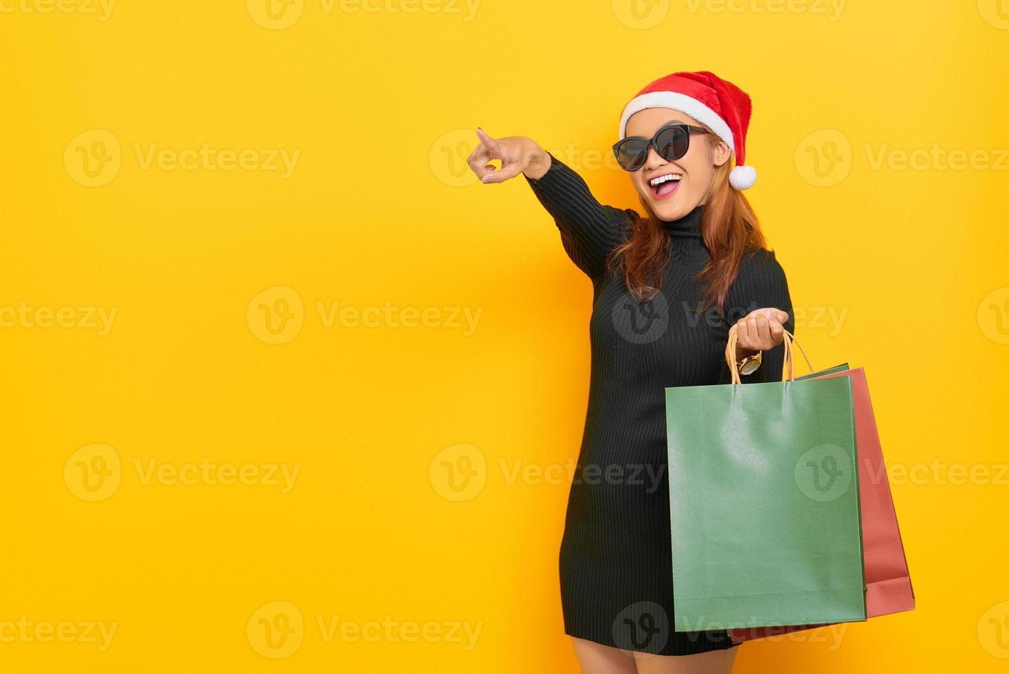 alegre jovem asiática com chapéu de Papai Noel e óculos de sol segurando sacolas de compras, apontando o dedo para o espaço da cópia isolado sobre fundo amarelo foto