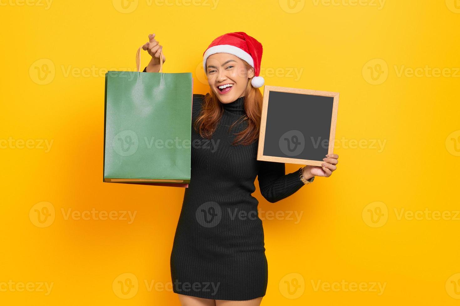 Mulher asiática jovem sorridente com chapéu de Papai Noel segurando sacolas de compras e um quadro-negro isolado sobre fundo amarelo foto