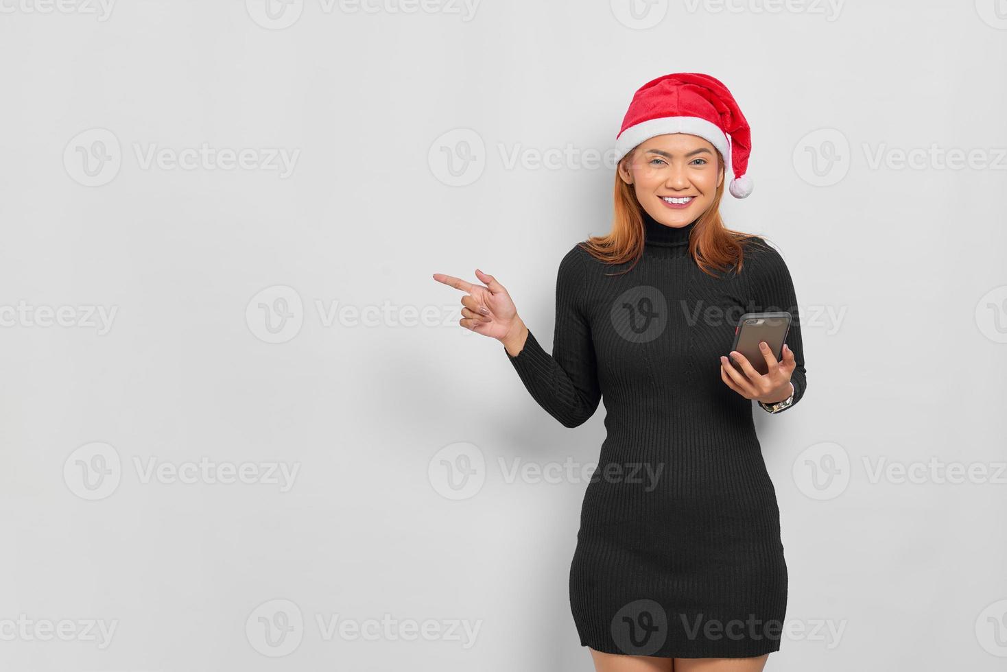 jovem asiática sorridente com chapéu de Papai Noel segurando um telefone celular e apontando o dedo para o espaço da cópia isolado sobre o fundo branco foto