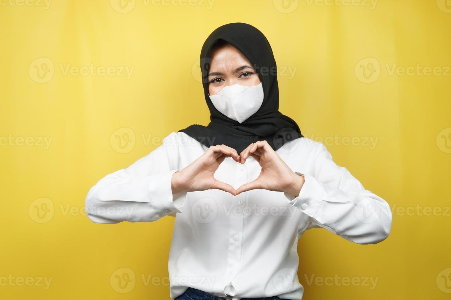 mulher muçulmana usando máscara branca, com sinal de mãos de amor, carinho, feliz, isolado em fundo amarelo foto