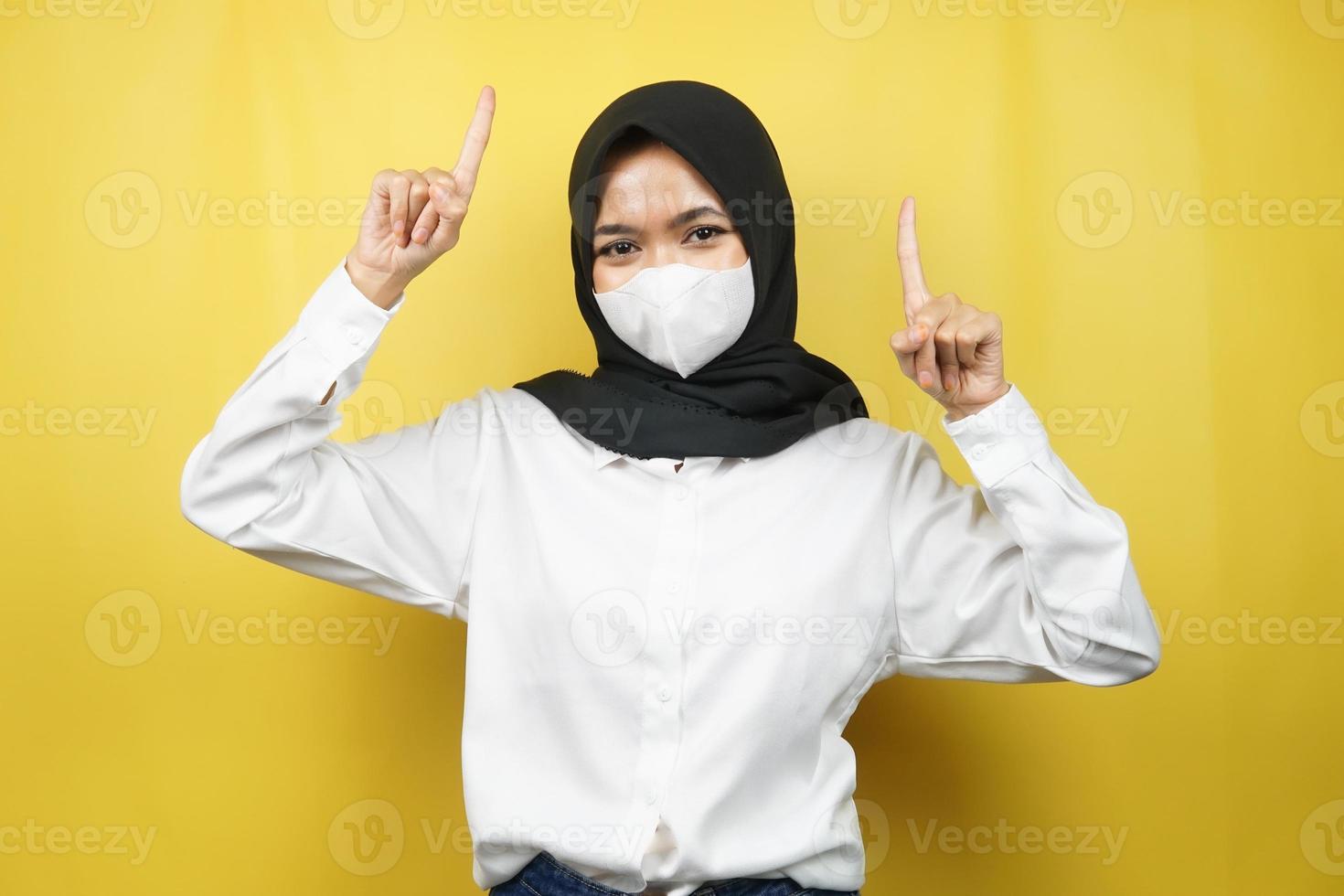 mulher muçulmana usando máscara branca, mão apontando para o espaço vazio, mão apontando para cima apresentando algo, isolado em fundo amarelo foto