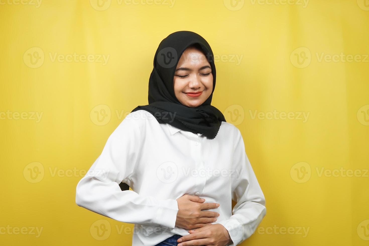 bela jovem asiática muçulmana sorrindo alegremente, com as mãos segurando a barriga, barriga cheia, isolada em fundo amarelo foto