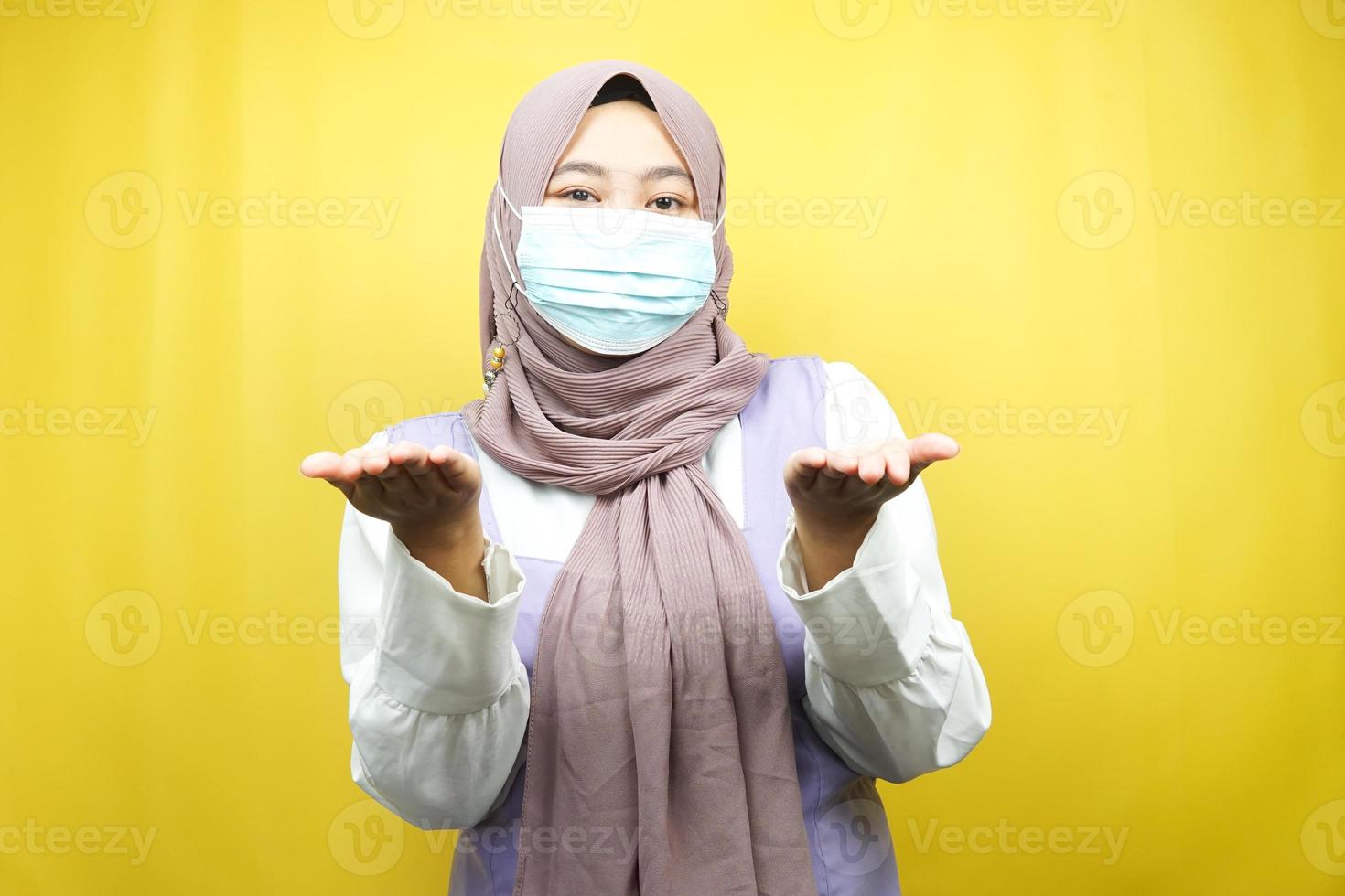 mulher muçulmana usando máscara médica, apresentando algo com as mãos, isolado em um fundo amarelo foto