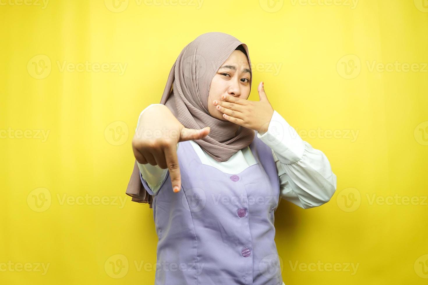 bela jovem muçulmana com a mão cobrindo a boca, a mão apontando para baixo, mostrando expressão de desaprovação, mostrando repulsa por algo, isolado em fundo amarelo foto