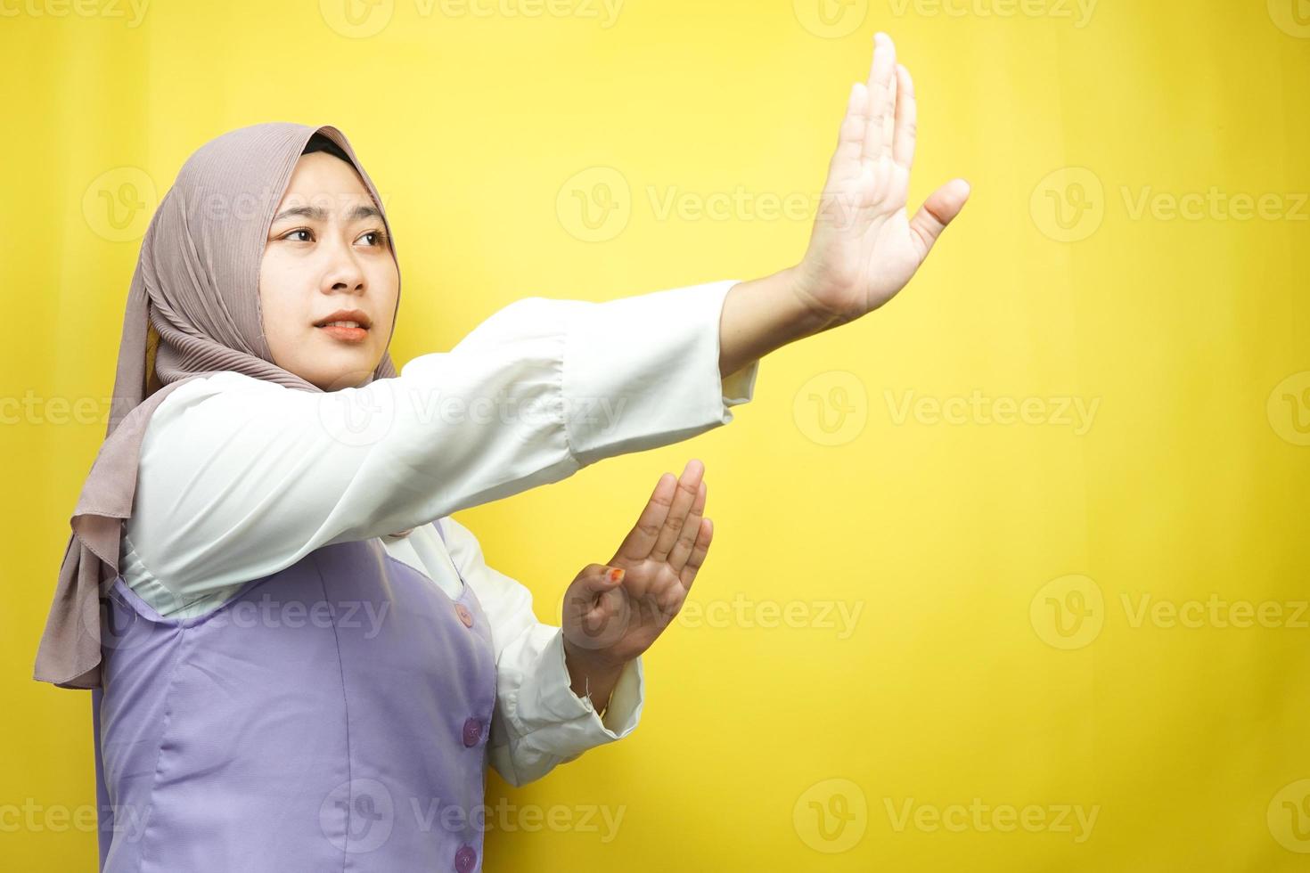 bela jovem muçulmana asiática com a mão rejeitando algo, a mão parando algo, não gostando de algo no espaço vazio, isolado no fundo amarelo foto