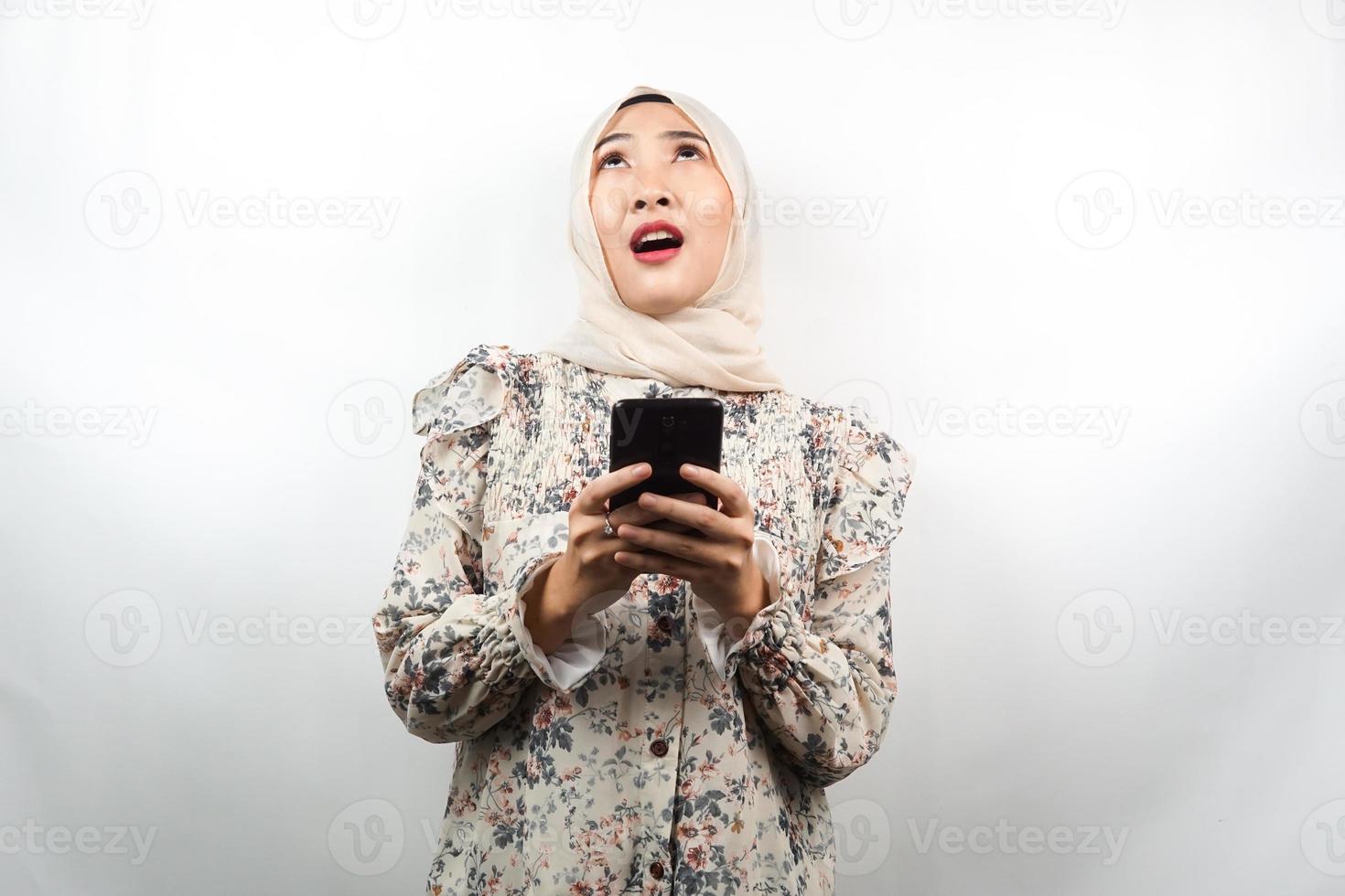 bela jovem muçulmana asiática chocada, surpresa, expressão uau, mão segurando o smartphone, olhando para cima, isolado no fundo branco foto