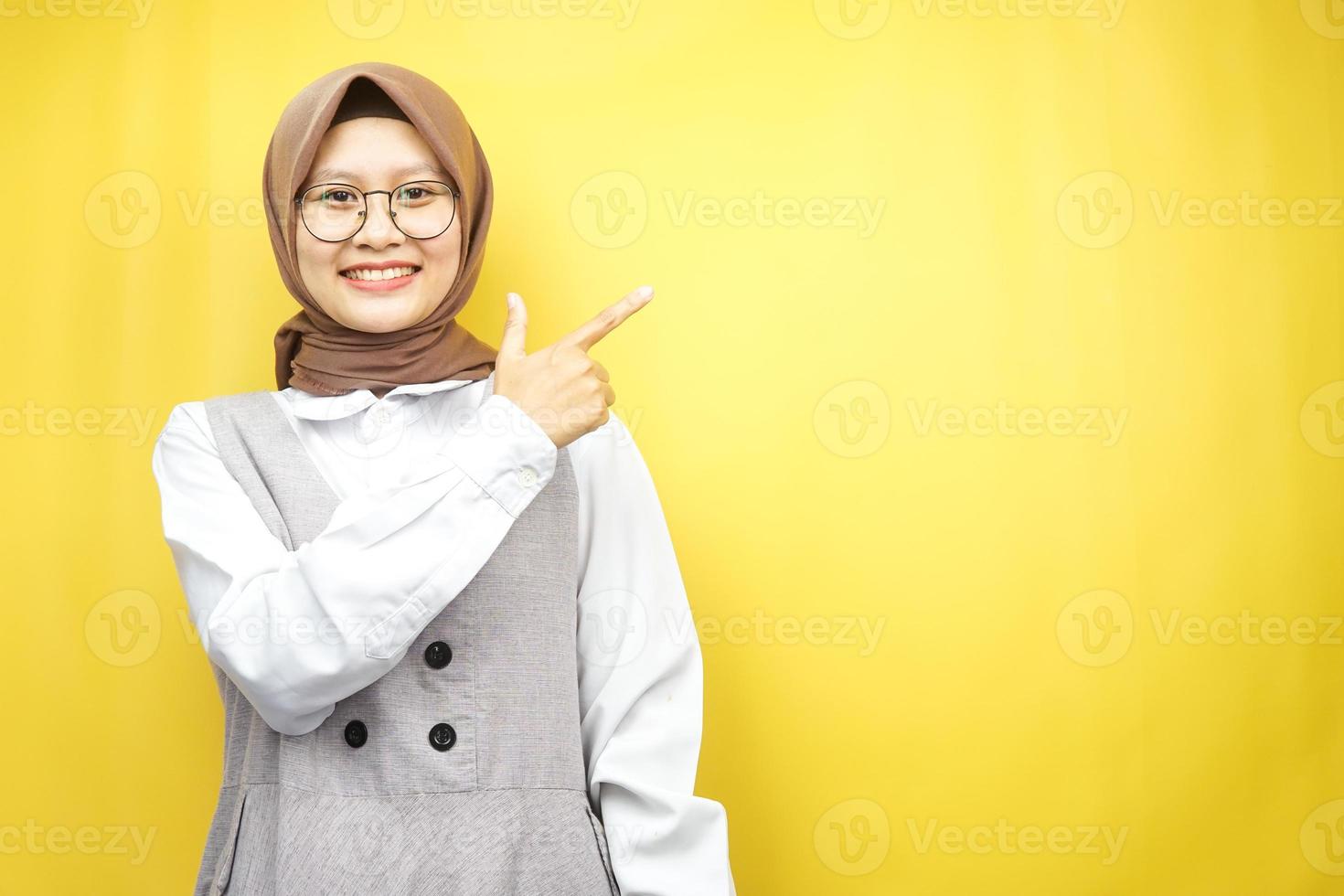 bela jovem asiática muçulmana com as mãos apontando o espaço vazio, apresentando algo, sorrindo confiante, entusiasmado, alegre, olhando para a câmera, isolado no fundo amarelo foto