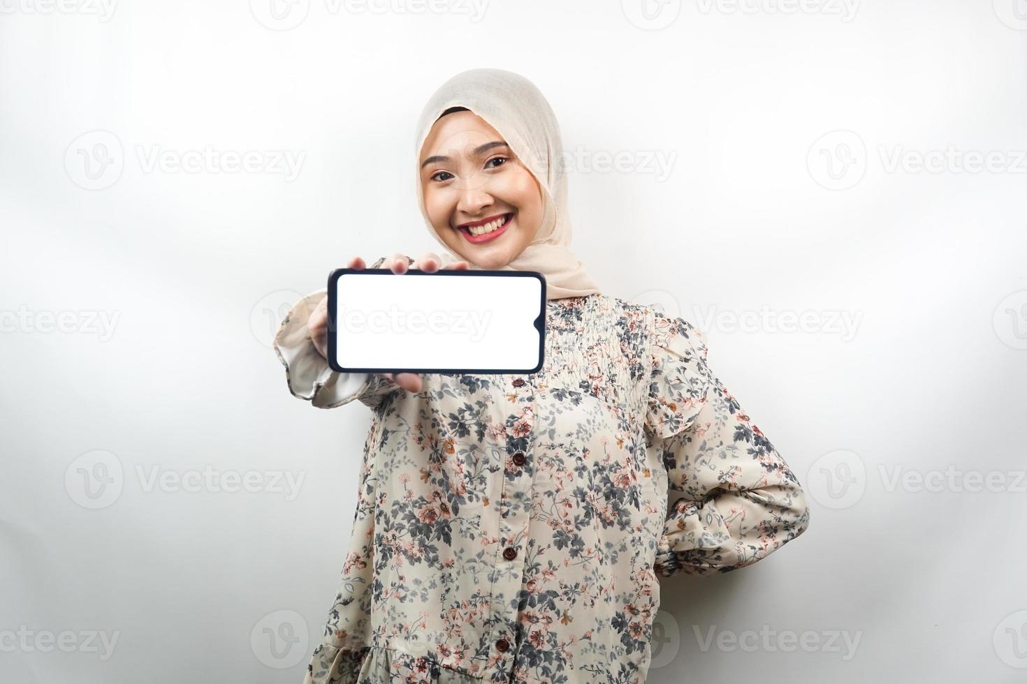 bela jovem muçulmana asiática sorrindo confiante, entusiasmada e alegre com as mãos segurando um smartphone, promovendo o aplicativo, promovendo o jogo, isolado no fundo branco, conceito de publicidade foto