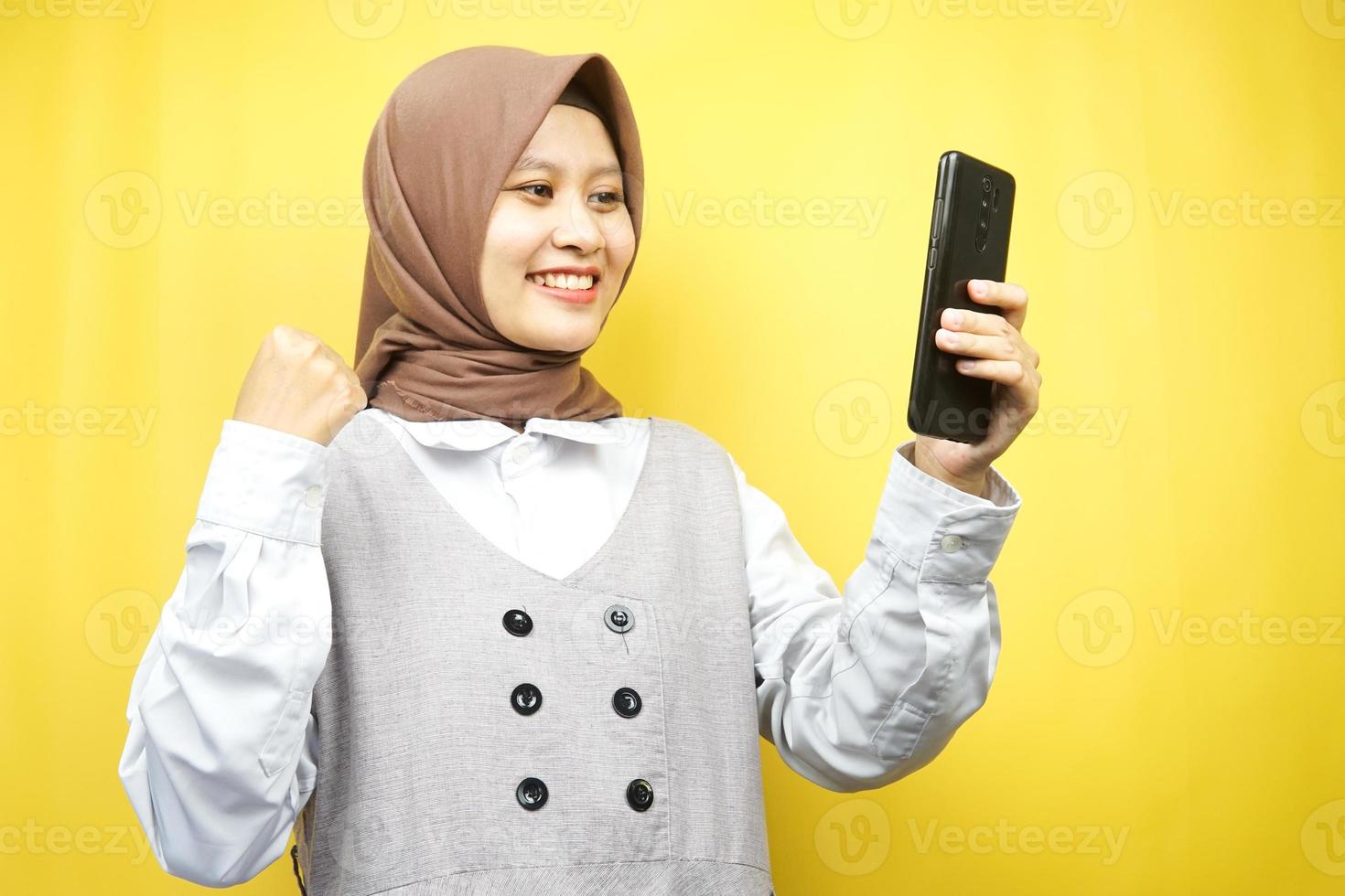 bela jovem muçulmana asiática sorrindo confiante, entusiasmada e alegre com as mãos segurando um smartphone, apertando as mãos, socando, vitória, sucesso, bom trabalho, isolado em fundo amarelo foto