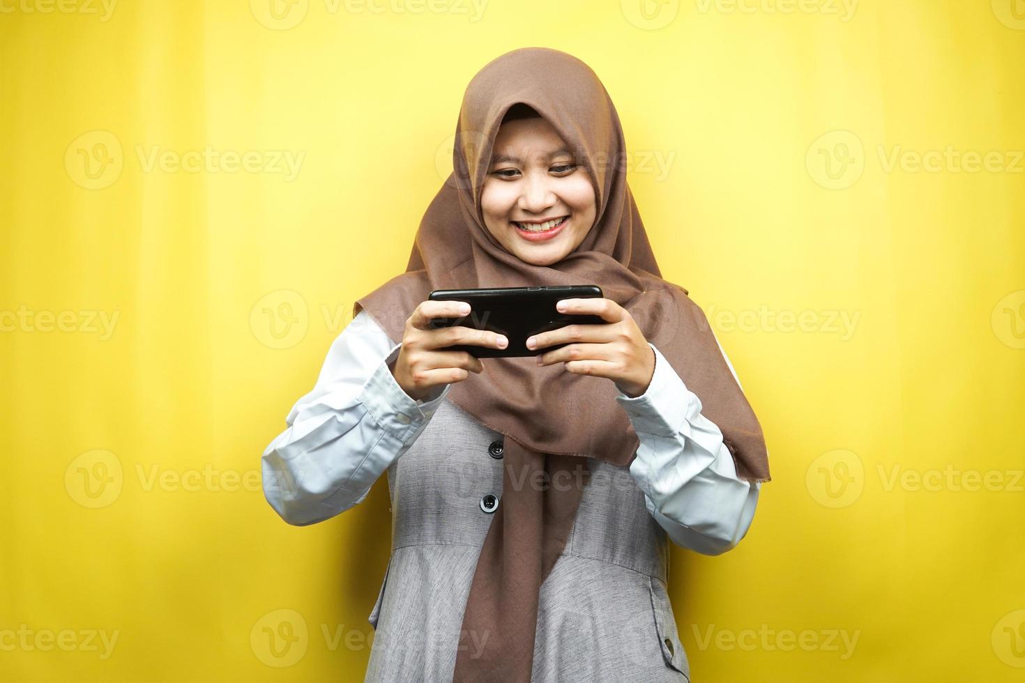 bela jovem muçulmana asiática com as mãos segurando um smartphone, jogando, sorrindo feliz, vitória, sucesso, isolado em um fundo amarelo, conceito de publicidade foto