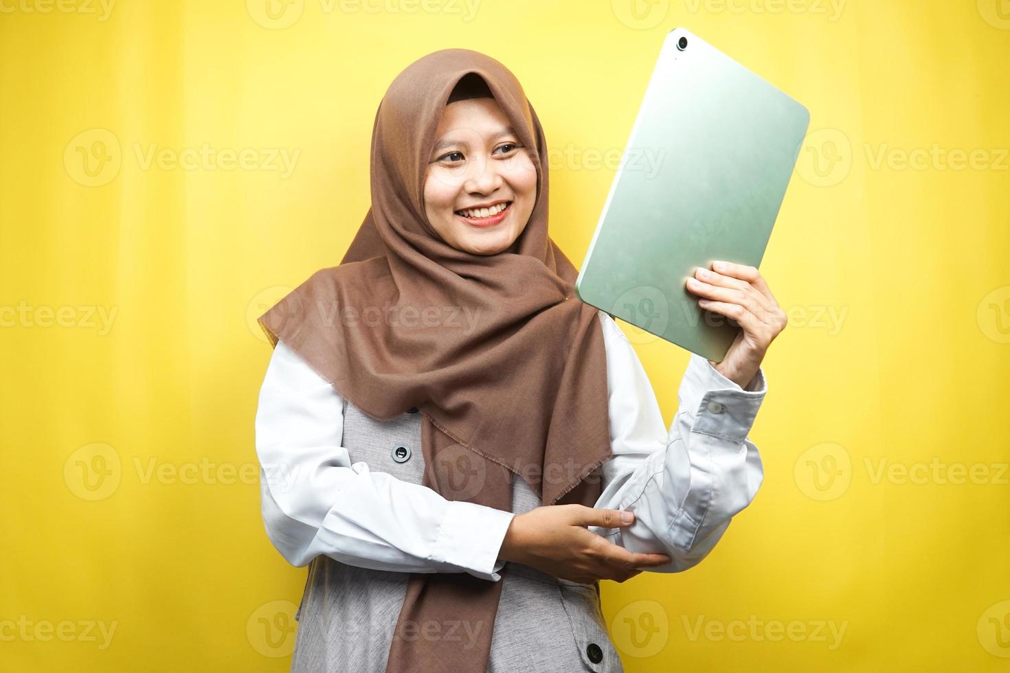 bela jovem asiática muçulmana sorrindo, animada e alegre segurando o tablet, isolado em um fundo amarelo foto