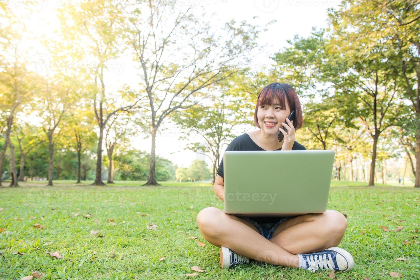 linda mulher asiática sorri e falando no celular enquanto está sentado no parque, dia de primavera. mulher asiática usando smart phone e laptop com sensação de relaxamento e rosto sorridente. conceitos de estilo de vida e tecnologia. foto