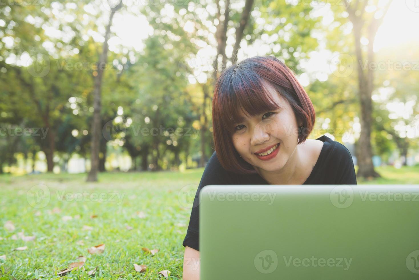 as pernas da jovem mulher asiática na grama verde com o laptop aberto. as mãos da menina no teclado. conceito de ensino à distância. hipster feliz jovem mulher asiática trabalhando no laptop no parque. estudante estudando ao ar livre. foto