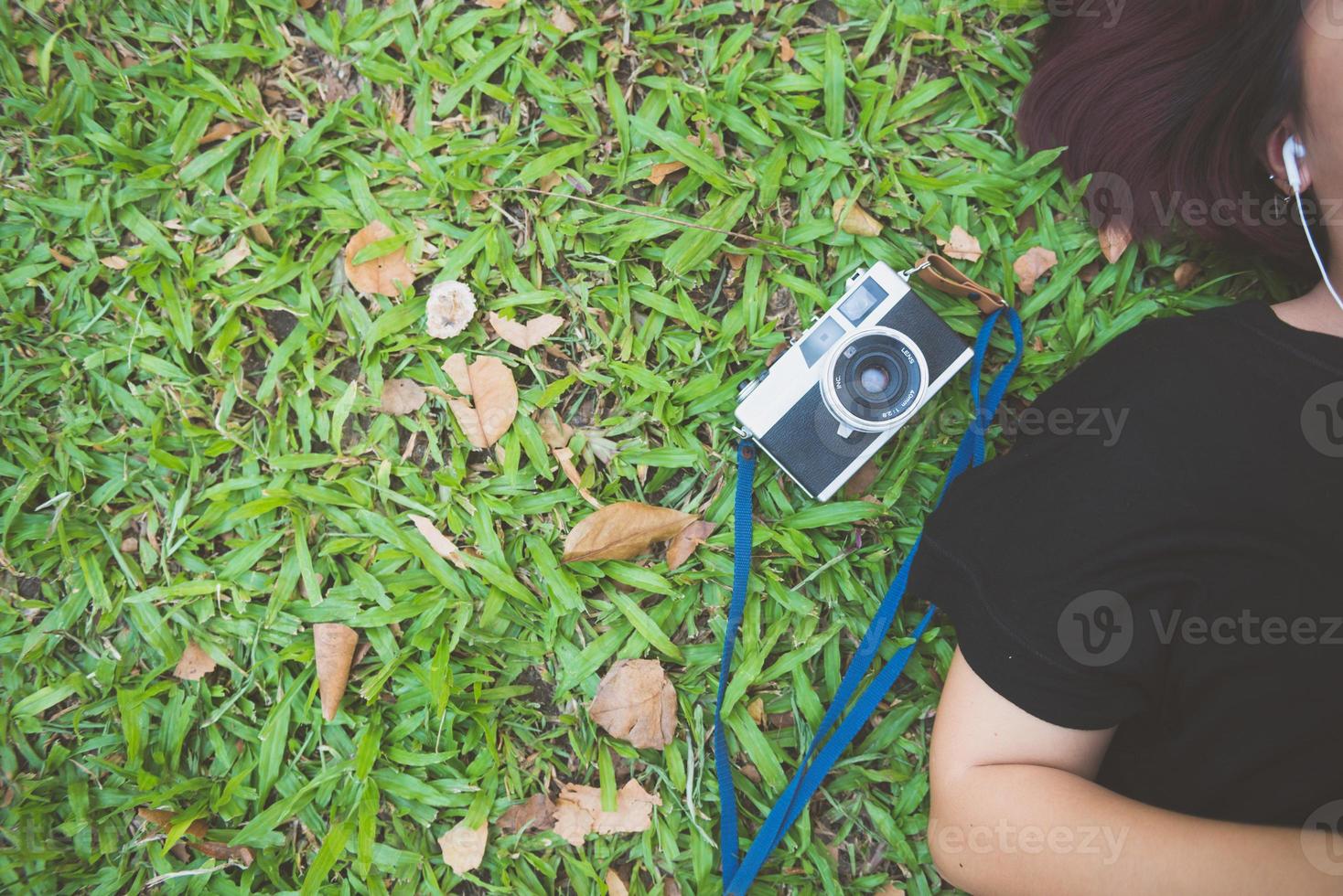 jovem mulher asiática deitado na grama verde, ouvindo música no parque, com uma emoção fria. jovem relaxando na grama com a câmera ao lado. atividade ao ar livre no conceito de parque. foto