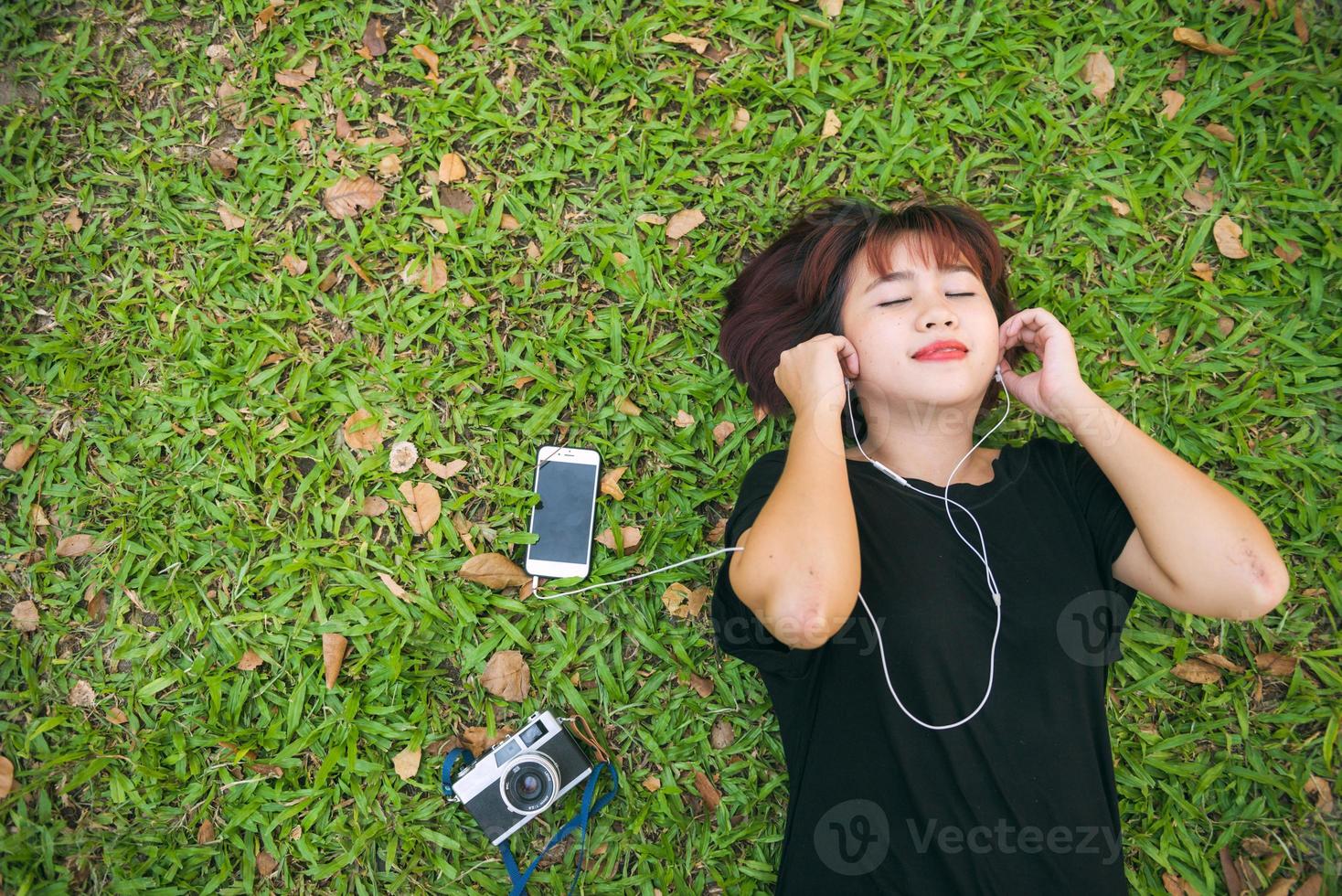 jovem mulher asiática deitado na grama verde, ouvindo música no parque, com uma emoção fria. jovem relaxando na grama com sua lista de reprodução de música. atividade ao ar livre no conceito de parque. foto