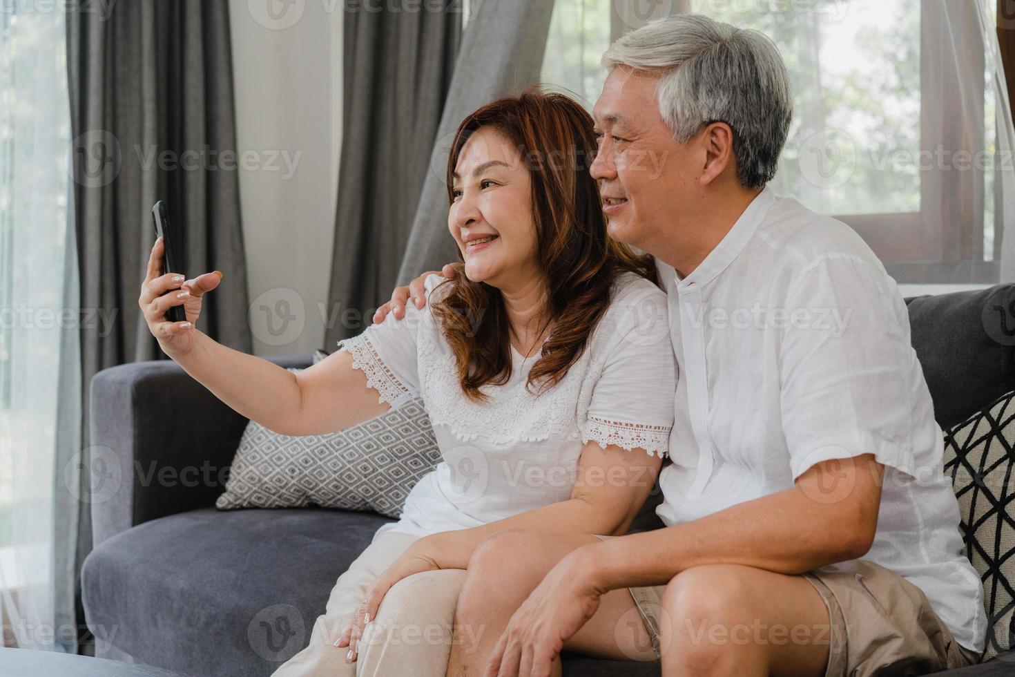 videochamada do casal sênior asiático em casa. avós chineses sênior asiáticos, usando a videochamada do telefone móvel, falando com os filhos dos netos da família, enquanto estava deitado no sofá na sala de estar em casa conceito. foto