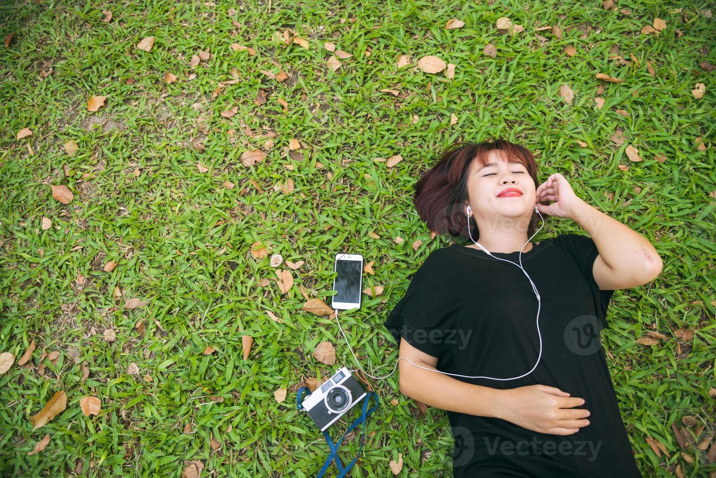 jovem mulher asiática deitado na grama verde, ouvindo música no parque, com uma emoção fria. jovem relaxando na grama com sua lista de reprodução de música. atividade ao ar livre no conceito de parque. foto