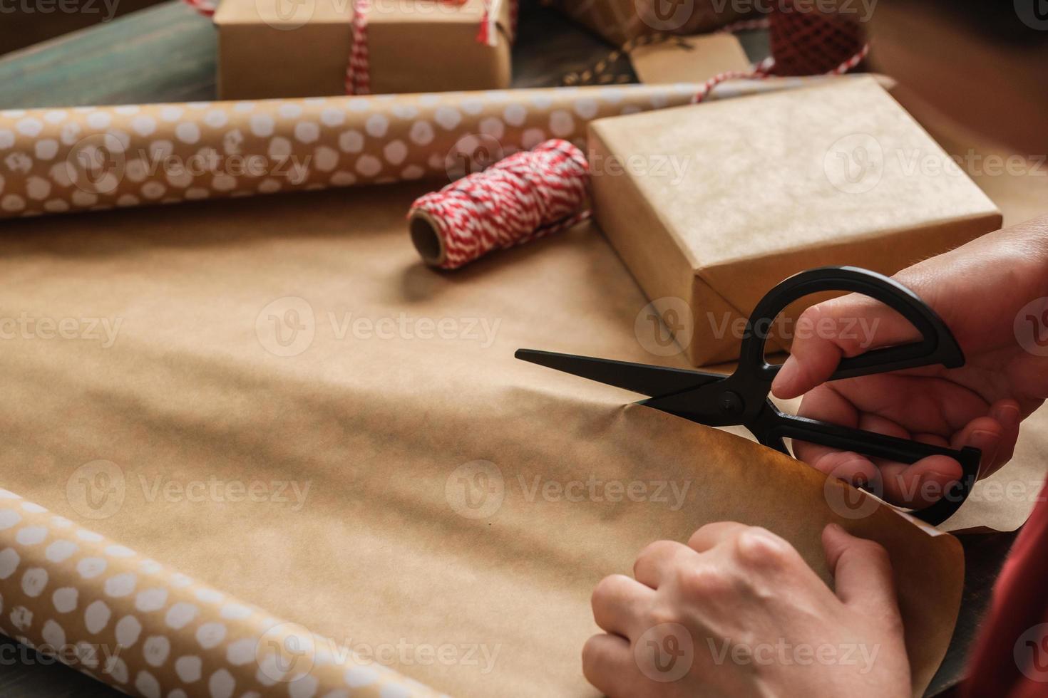 Feche a mão da mulher cortando papel kraft para embrulhar a caixa de presente de Natal na mesa de madeira foto