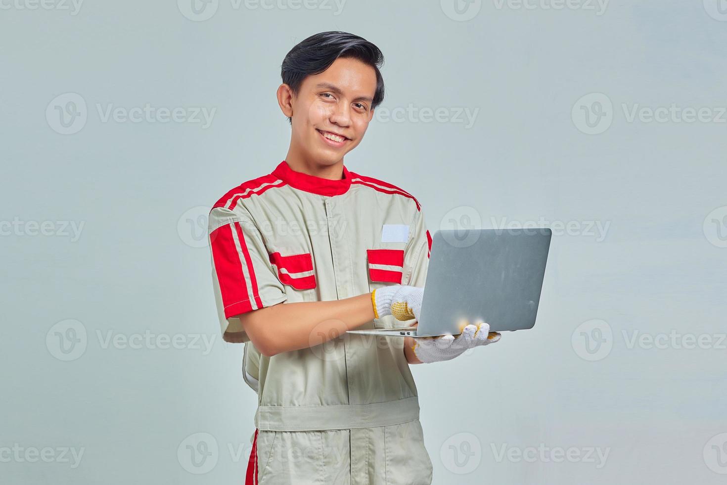 retrato de um homem bonito e sorridente mecânico vestindo uniforme segurando e usando laptop em fundo cinza foto